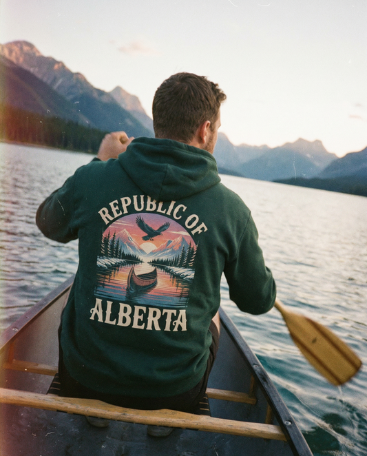Person in a canoe on a lake with mountains in the background, wearing a hoodie with 'Republic of Alberta' design.
