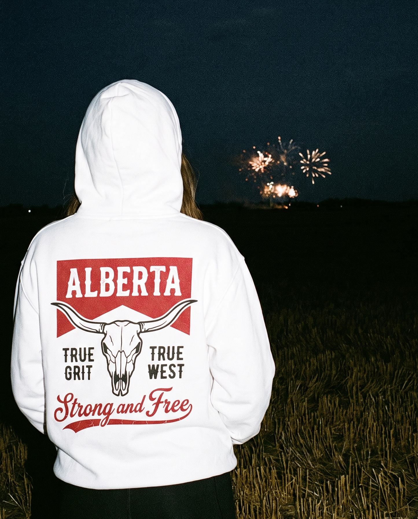 A person viewed from behind wearing a white pullover hoodie with a large Alberta-themed back graphic featuring a longhorn skull, red and black text, and fireworks visible in the night sky above a field.