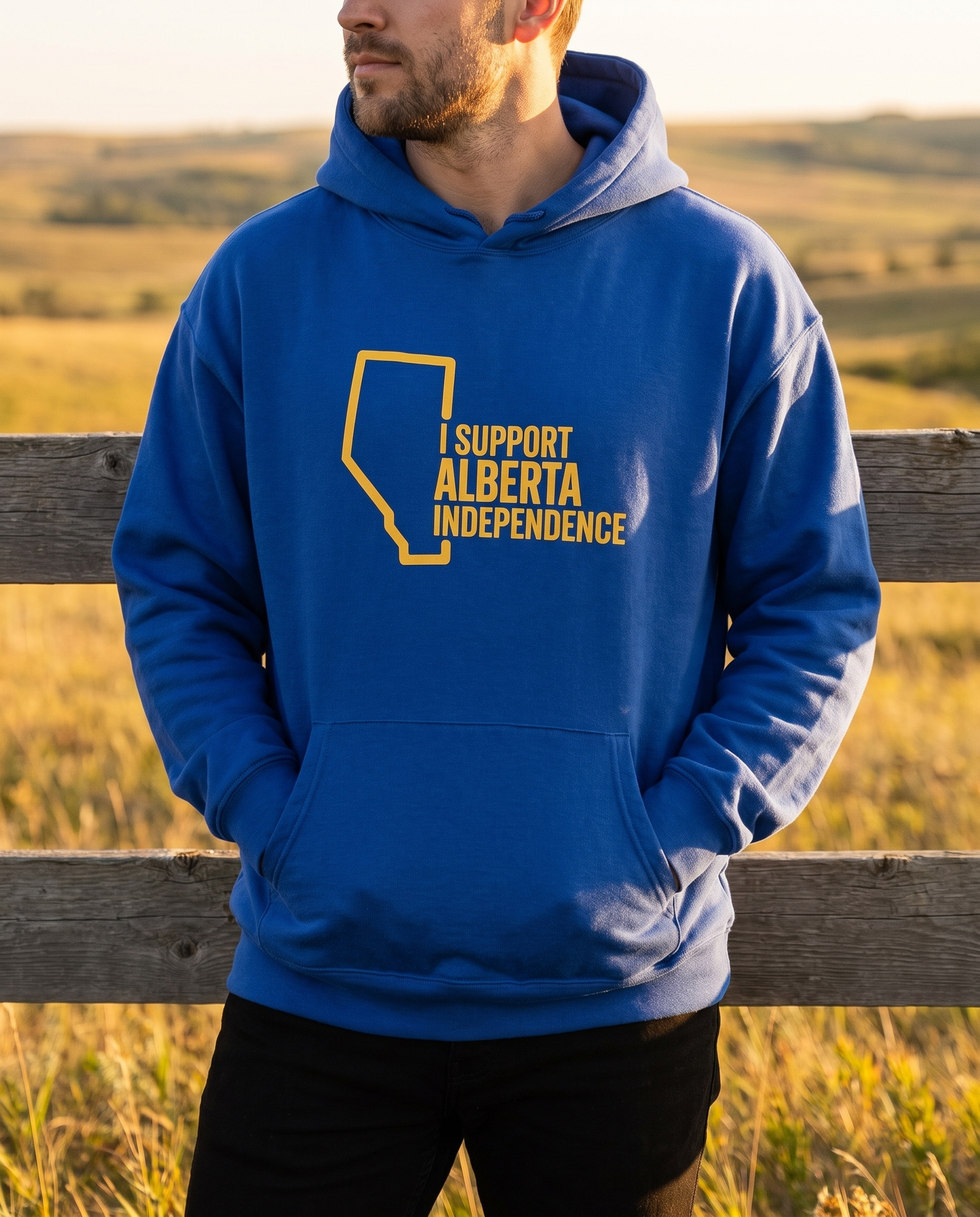 Man wearing cobalt Support Alberta Independence oversized hoodie at ranch fence golden hour