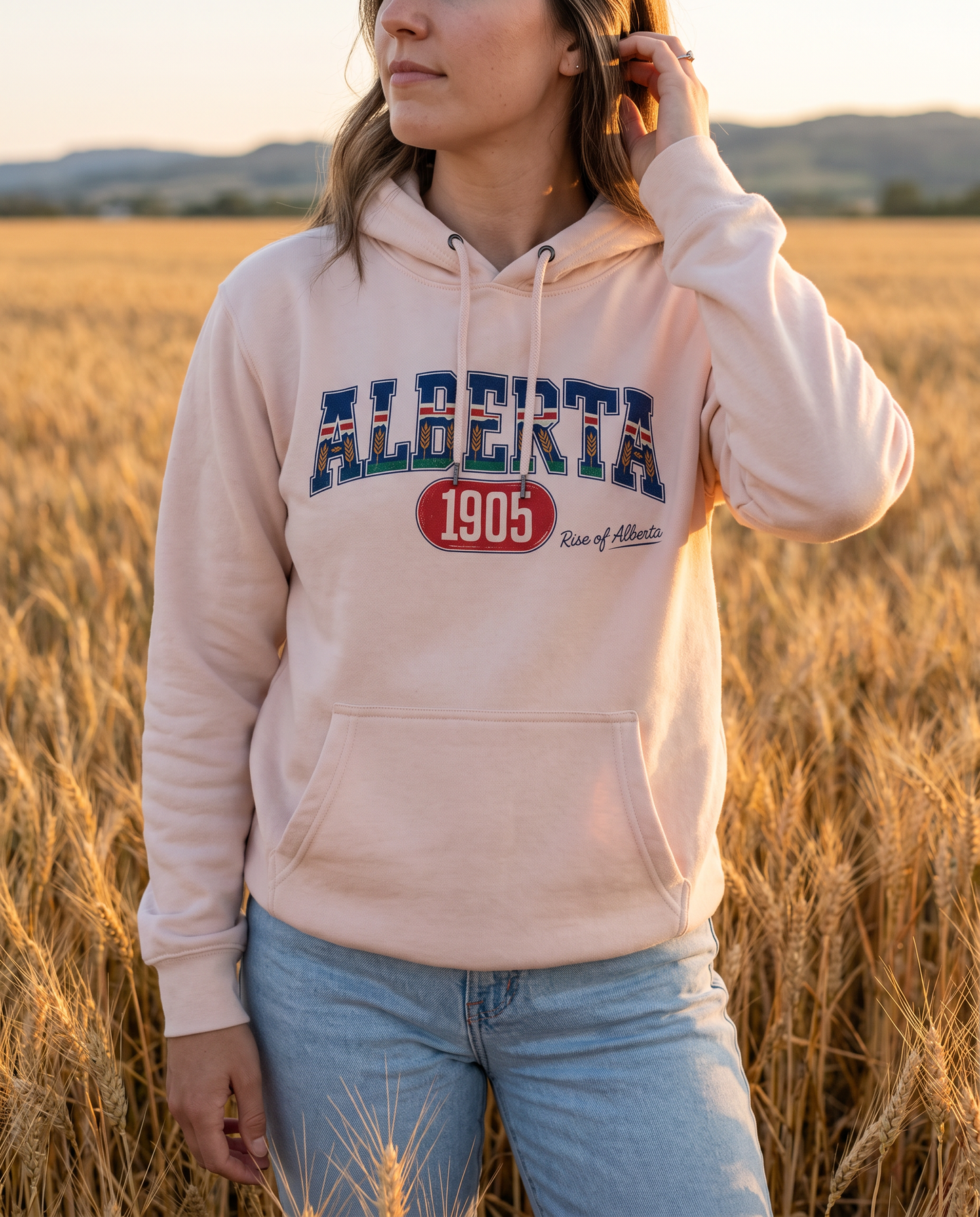 Woman wearing pale pink Alberta 1905 hoodie in wheat field at golden hour