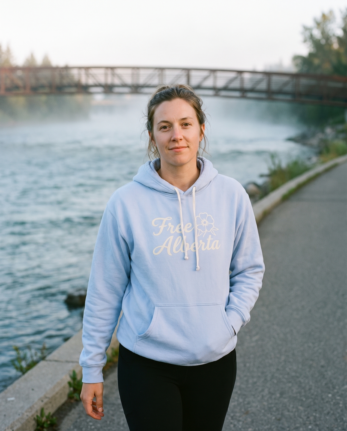 Woman wearing the Hydrangea Free Alberta hoodie on a river pathway at dawn
